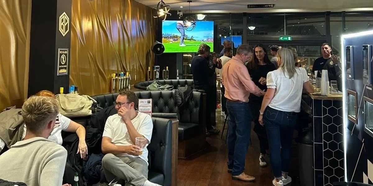 Guests relaxing in the sports bar at Ball Park Brighton Marina, with black leather booths, gold wall panels and a TV showing golf
