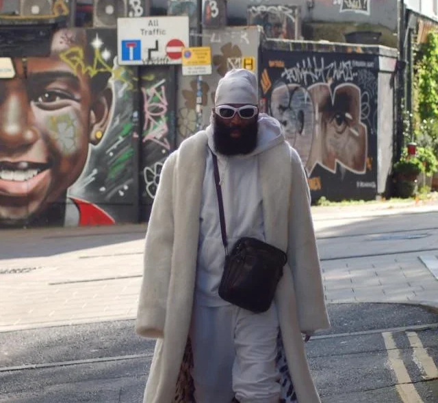 Charles "Pirate" Saddler in an all-white fur coat and grey tracksuit with sunglasses, standing on a Brighton street corner in front of graffiti murals