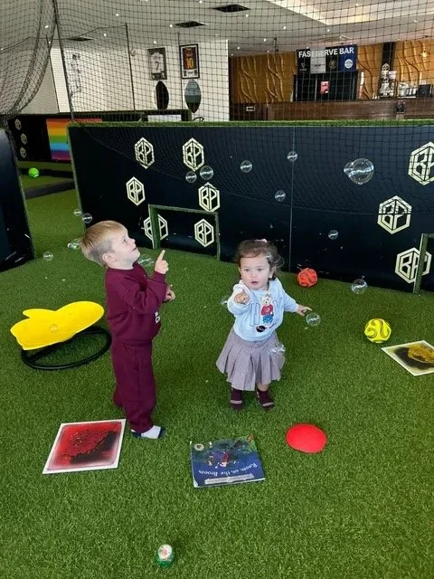 Two toddlers pointing up at bubbles on the artificial turf at Ball Park Brighton Marina, with soft footballs, a yellow rocker, sensory light mats and a picture book on the floor and the Fast Serve Bar behind them