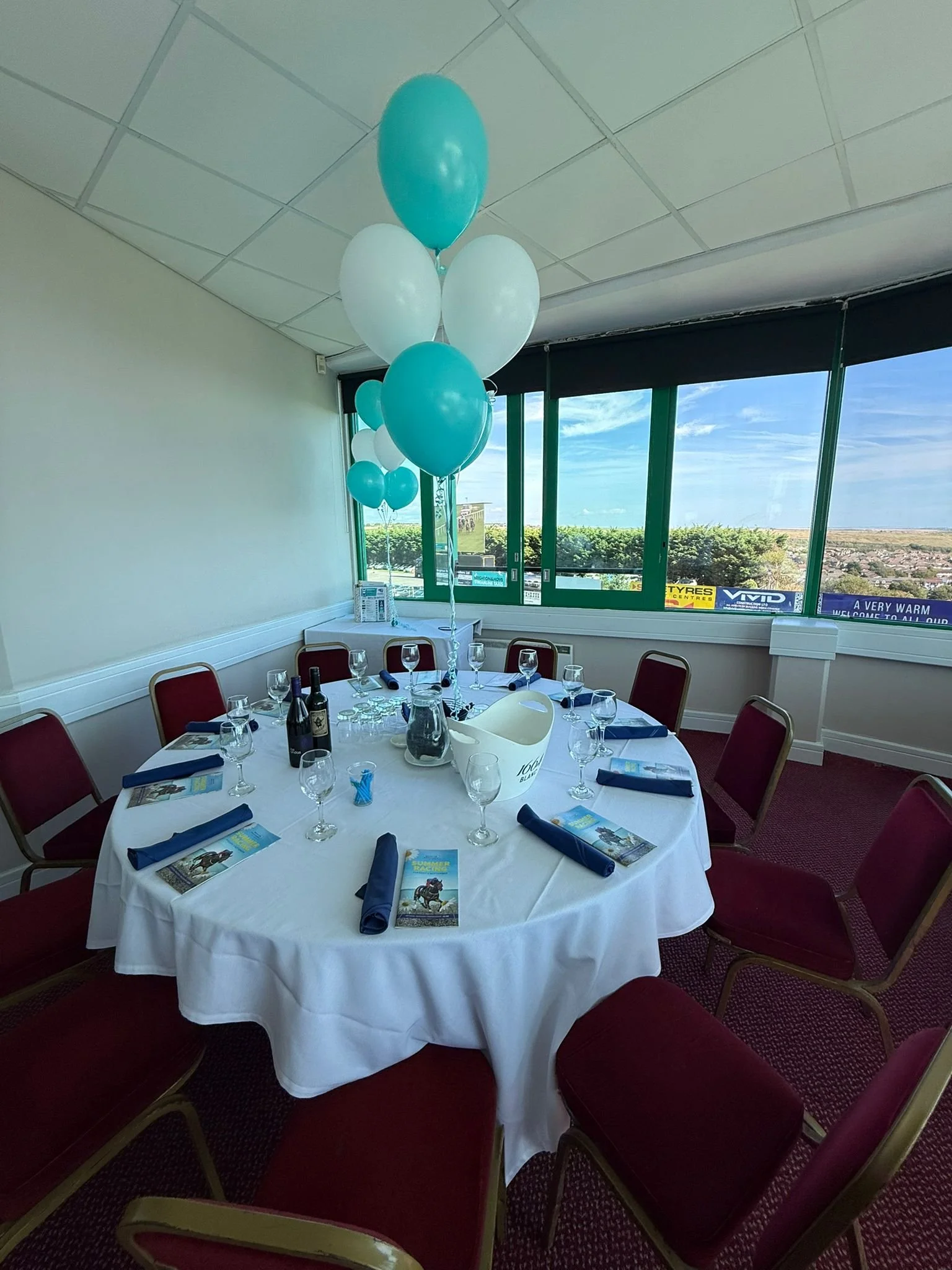 Table set with wine and racecards beneath teal and white balloons overlooking Brighton Racecourse