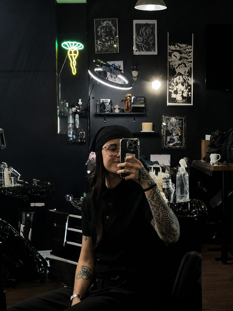 Copito, wearing black with a headscarf and glasses, takes a mirror selfie in a dark tattoo studio with neon signs and framed artwork on the walls