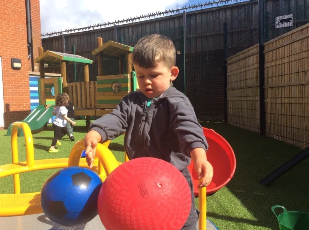 Toddler in a grey zip-up hoodie reaching for a blue football-pattern ball and a red playground ball in the outdoor play area at the Jeanne Saunders Centre SEND nursery in Hove, with a wooden climbing frame and slide behind