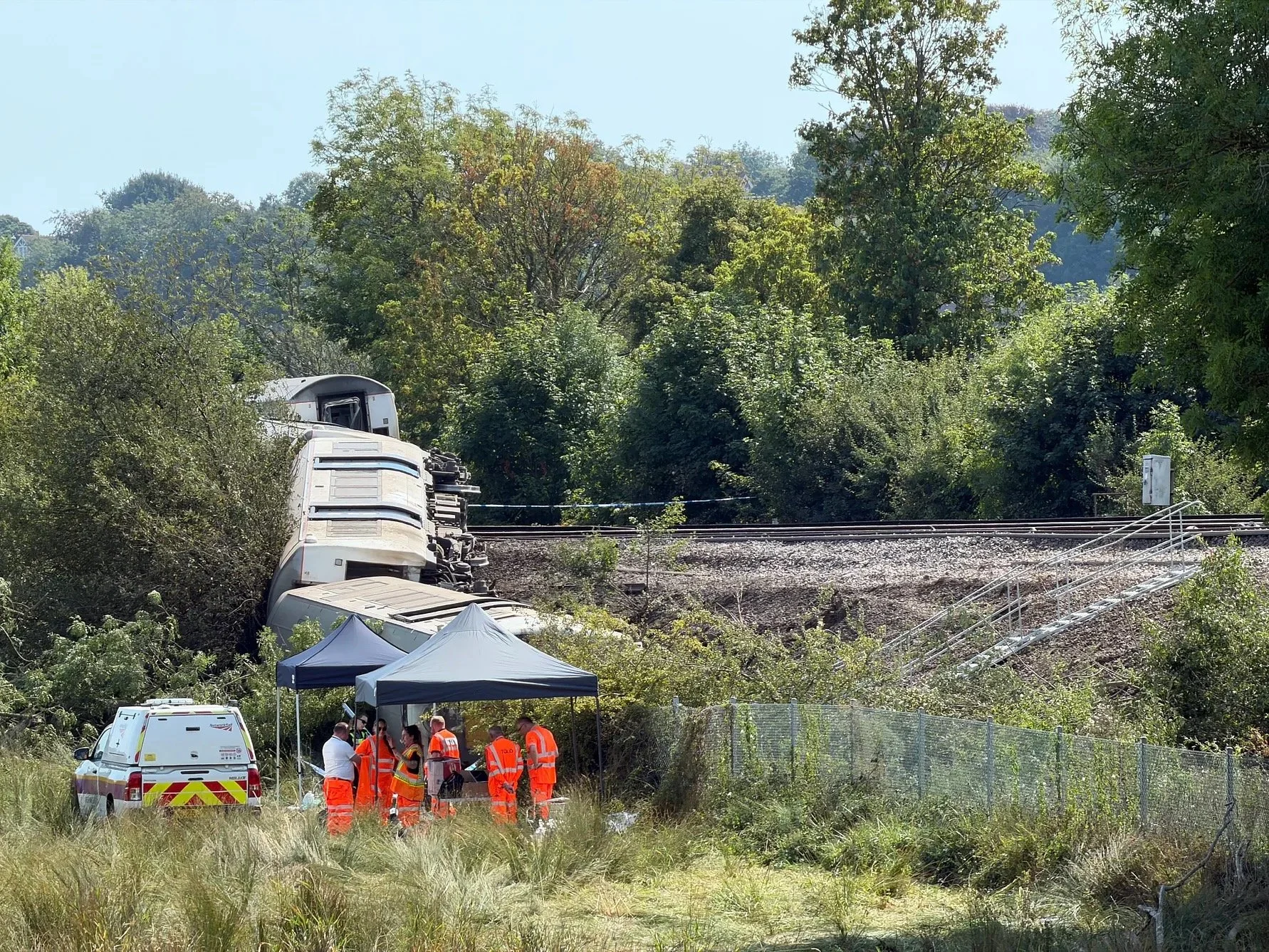 Alt: Overturned train carriages at the recovery site near Lewes with orange-clad Network Rail workers and a response vehicle beside the track