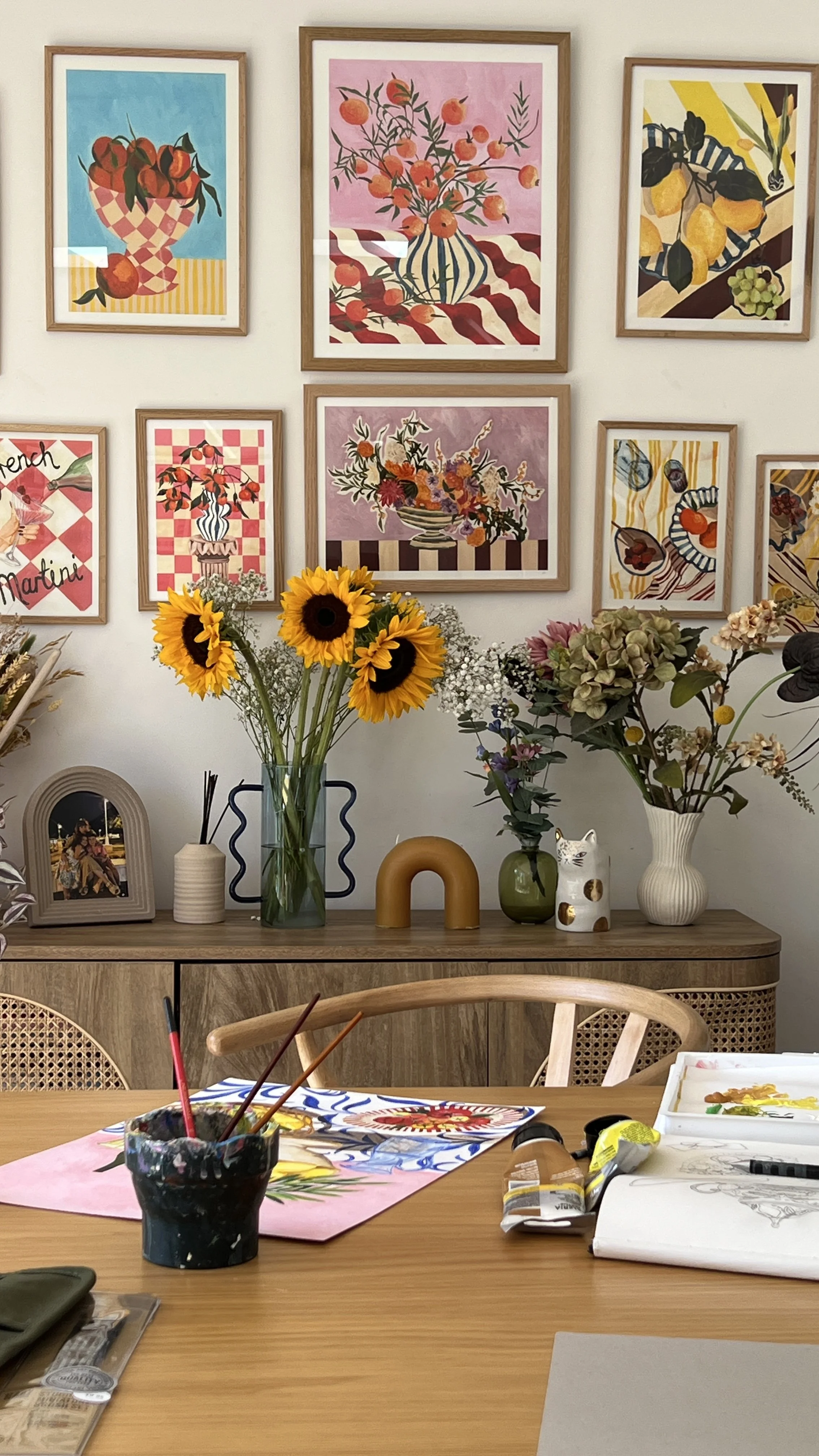 Hand-painted framed prints displayed on a studio wall at Violets Print House, featuring botanical and geometric designs in warm tones, with fresh flowers and design materials on the wooden desk below