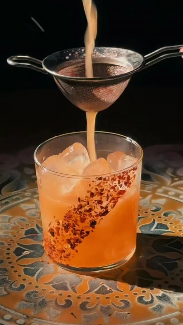 A cocktail with ice in a glass, with a filter and poured liquid being strained into it, on a decorative surface.