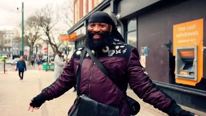 Charles "Pirate" Saddler laughing with arms outstretched on a Brighton street, wearing a purple jacket with badges and a black headscarf