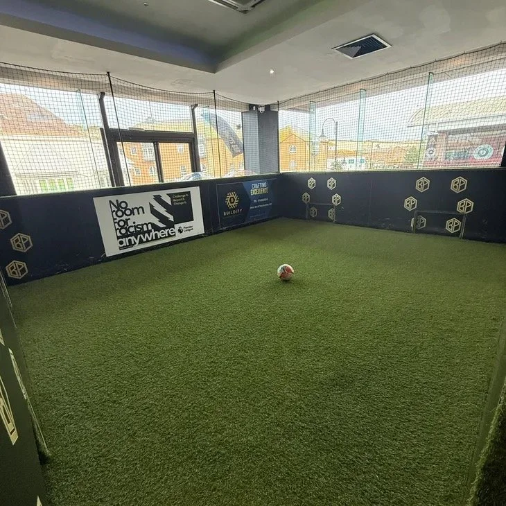 The Quick Pitch at Ball Park Brighton Marina, an enclosed indoor mini pitch with artificial turf, a football in the centre, small goals in the black perimeter wall and windows looking out over the marina