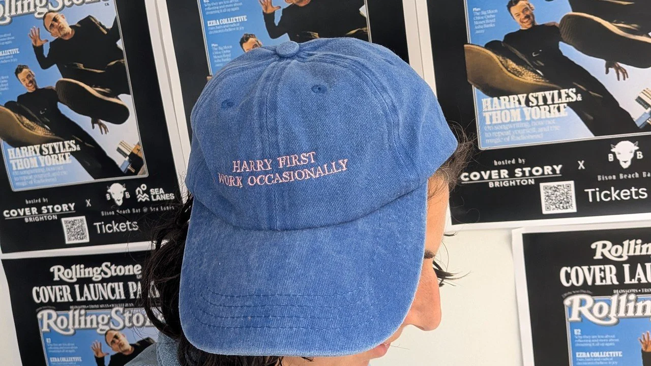 A blue cap embroidered with Harry first, work occasionally, in front of Rolling Stone UK cover launch party posters for the Harry Styles and Thom Yorke issue at Bison Beach Bar at Sea Lanes, Brighton
