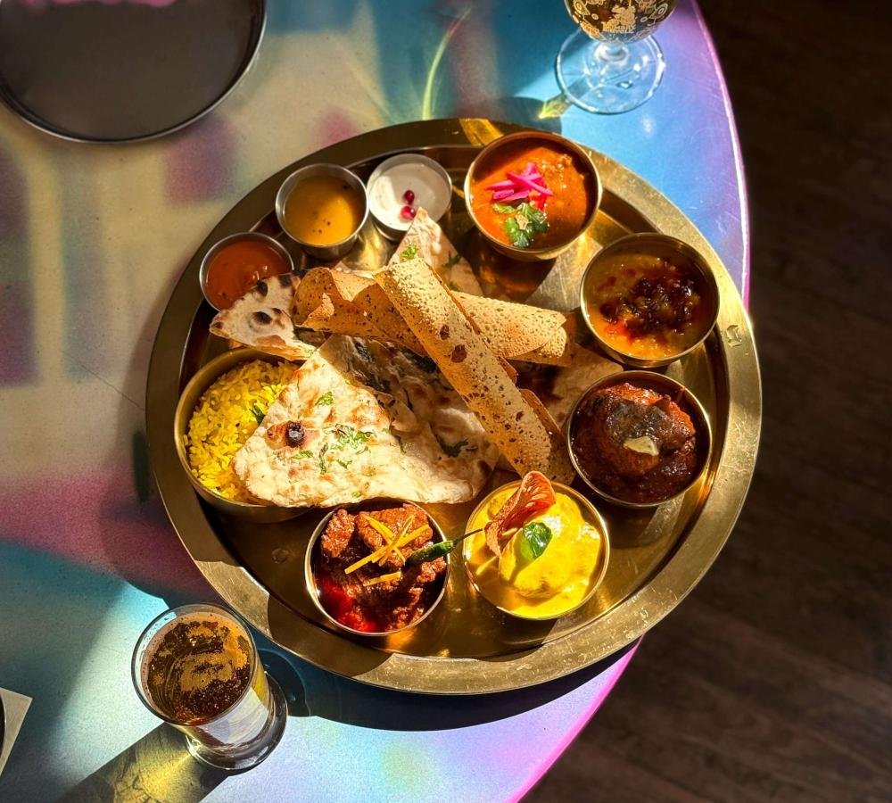 A traditional Indian thali on a round metal tray, including rice, naan bread, various curries, chutneys, and desserts, with a glass of brown drink and an empty glass nearby.