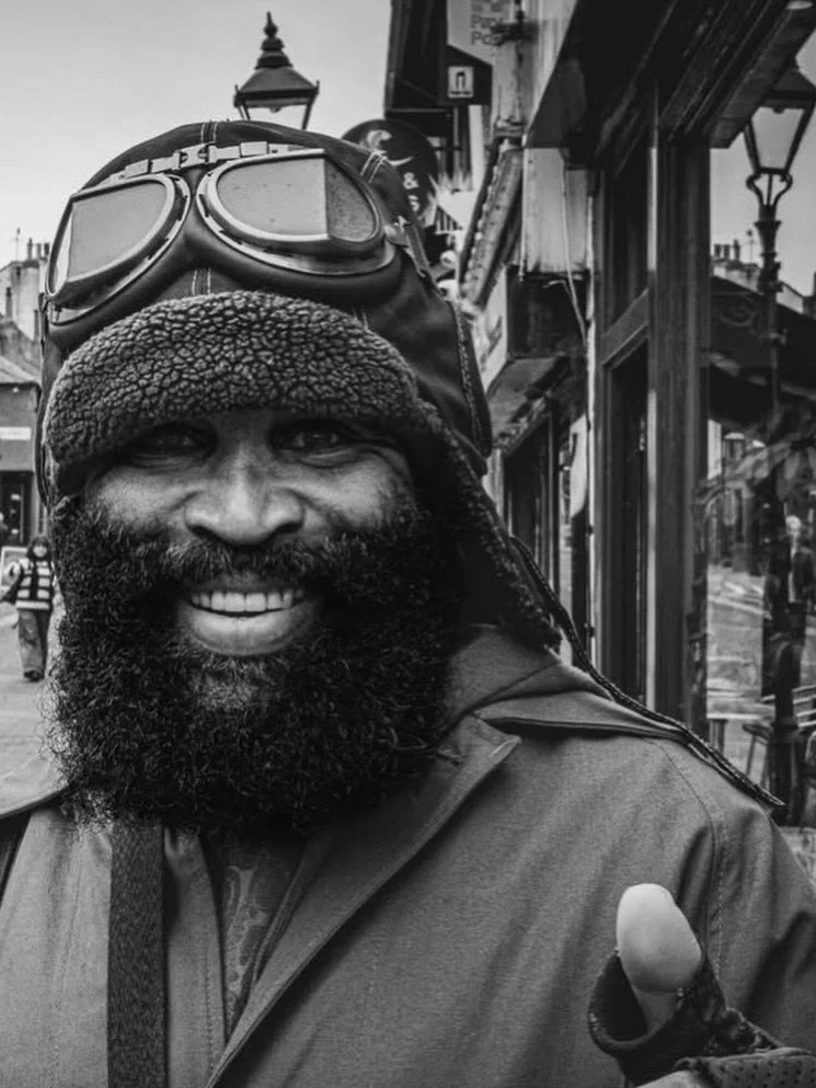 Black and white portrait of Charles Pirate Saddler smiling on a Brighton street in his aviator cap and goggles