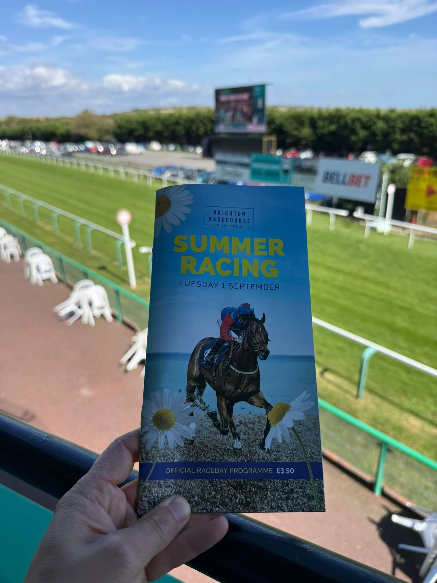 Hand holding the Brighton Racecourse Summer Racing programme for Tuesday 1 September with the track behind