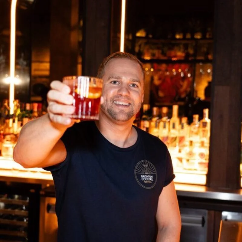 Brighton Cocktail Festival founder Harry Philippson raising a cocktail in a bar wearing a festival T-shirt