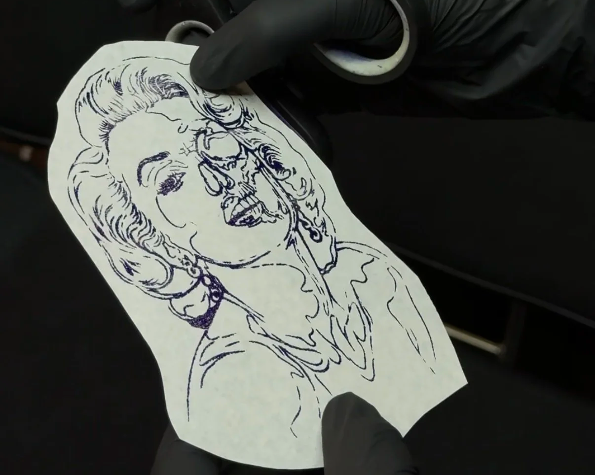A gloved hand and scissors cutting out the blue-ink line stencil of the Marilyn Monroe and skull design before tattooing