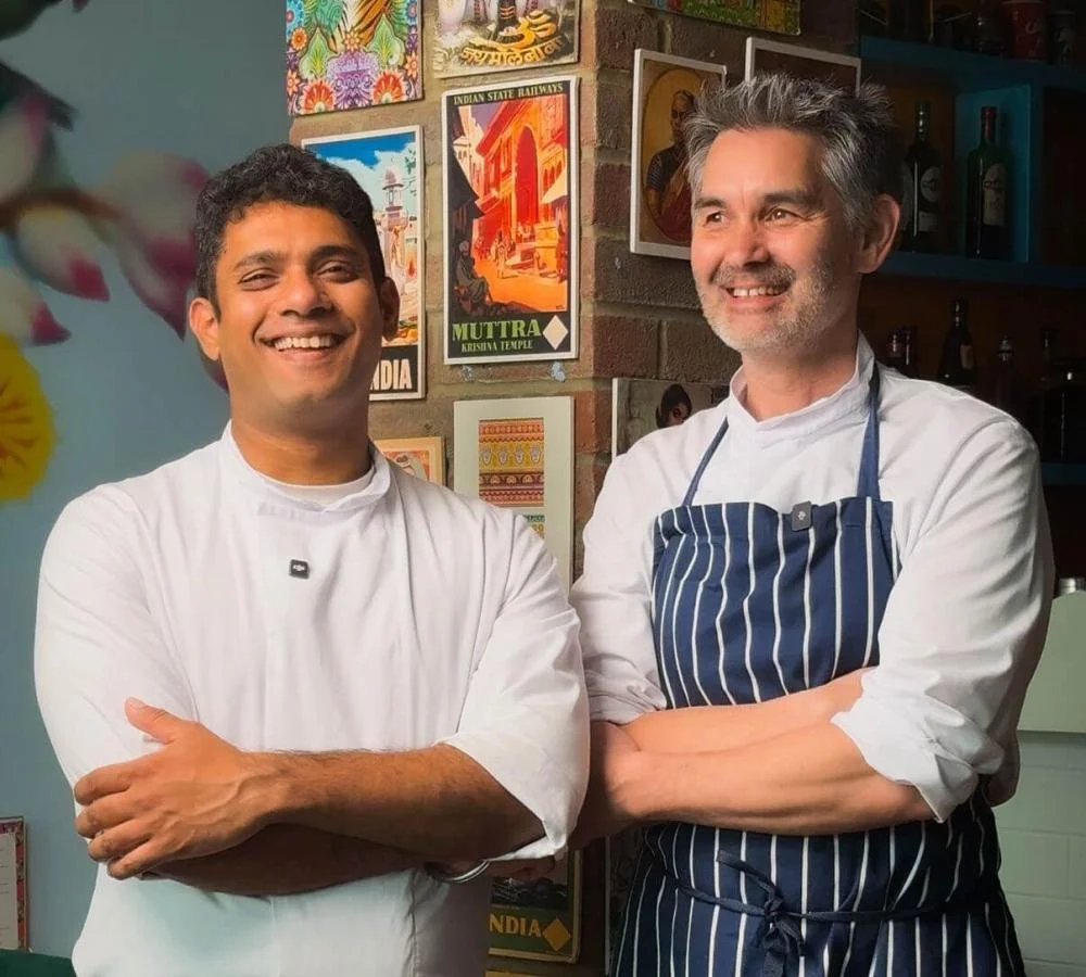 Two male chefs standing in a restaurant kitchen smiling with their arms crossed, one wearing a white chef's coat and the other wearing a white shirt with a blue and white striped apron, with colorful posters and bottles in the background.