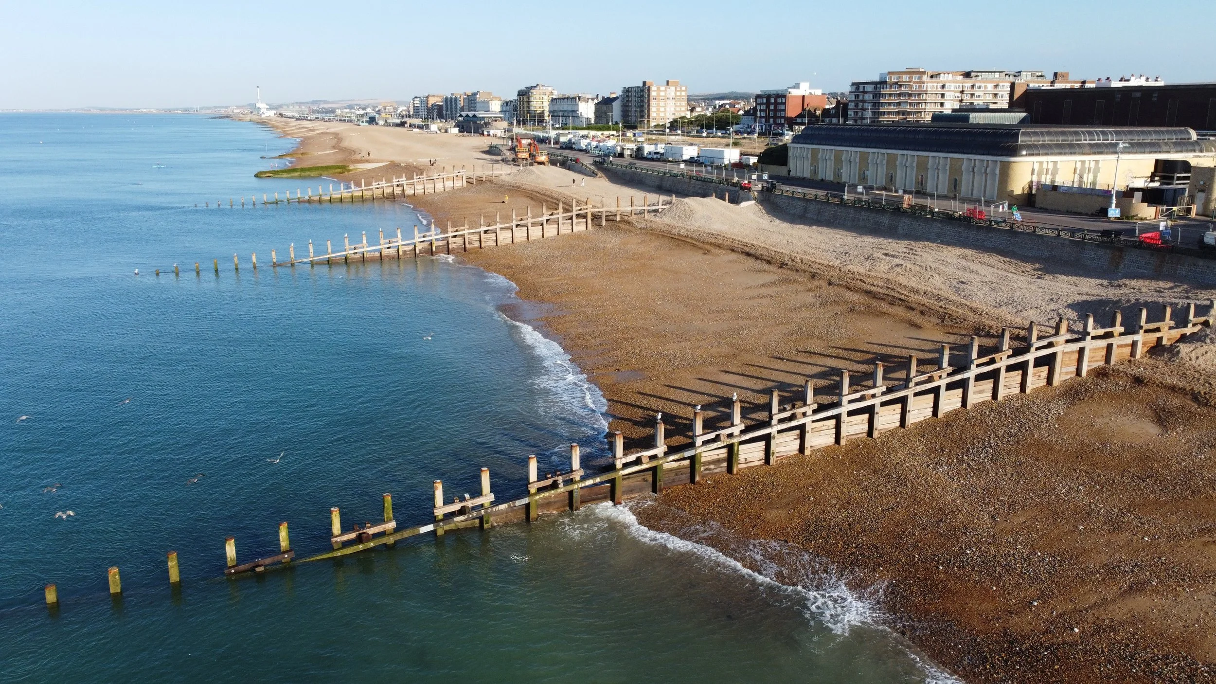 Aerial view of Hove beach at low sun showing timber groynes running down the shingle into a calm sea, with the promenade, King Alfred Leisure Centre and seafront blocks behind