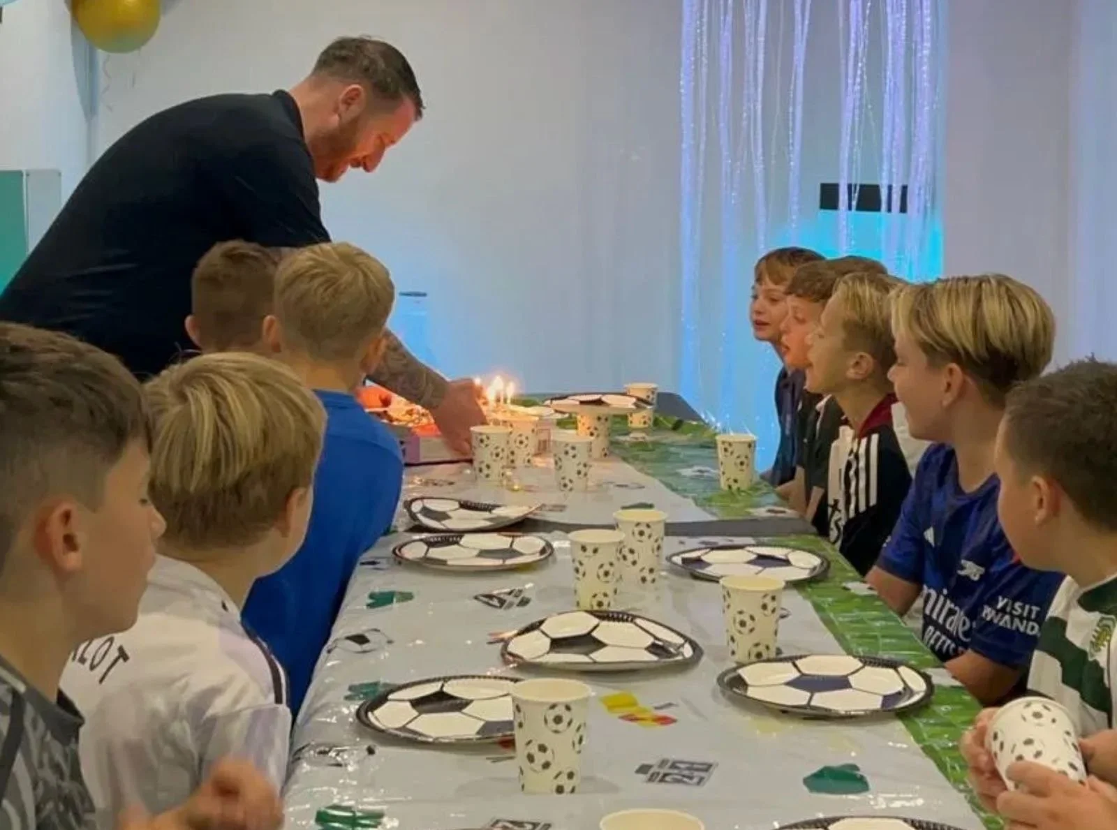 A man lighting candles on a birthday cake at a long party table set with football-themed plates and cups, while a row of young boys in football shirts watch, with a fibre optic light curtain glowing behind them
