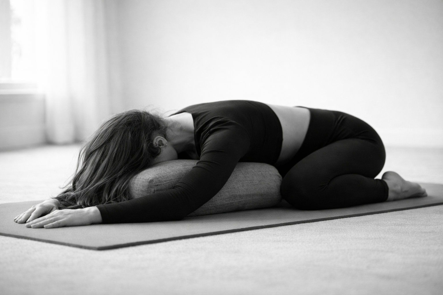 A woman practicing yoga on a mat, leaning forward with her arms extended and resting her head on a cushion in a bright room.