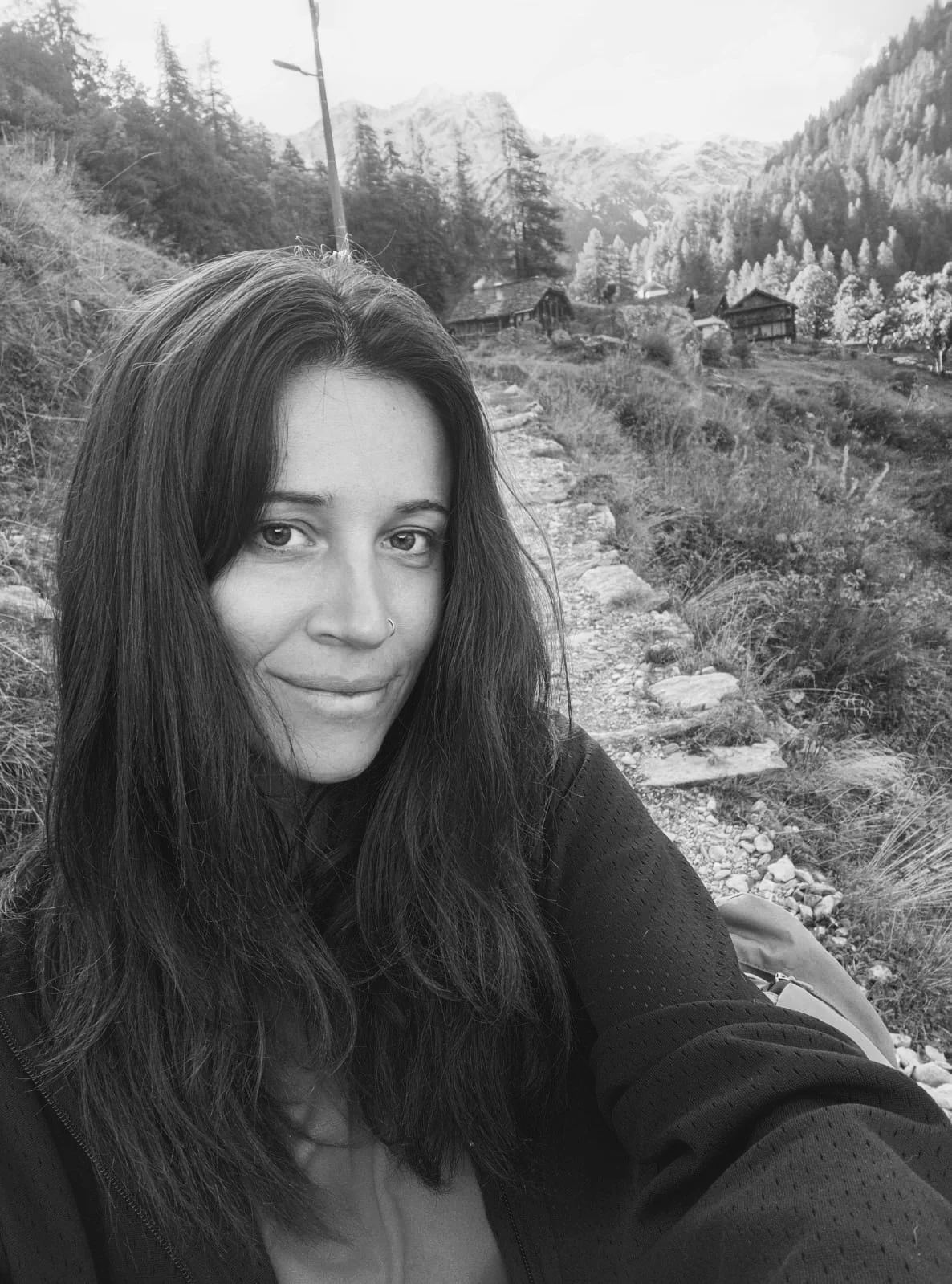 A woman with long dark hair, a nose piercing, and a slight smile taking a selfie outdoors on a mountain trail, with mountains and trees in the background.