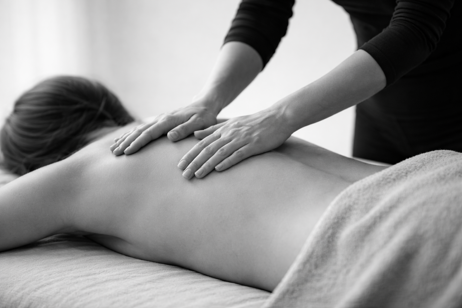 Black and white photo of a person receiving a back massage from a masseuse. The person is lying face down on a massage table, partially covered with a towel.