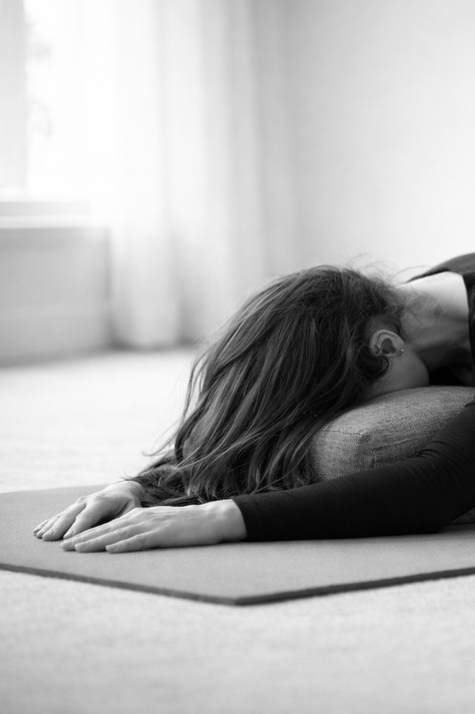 A woman practicing yoga, performing a child's pose on a mat in a bright room.