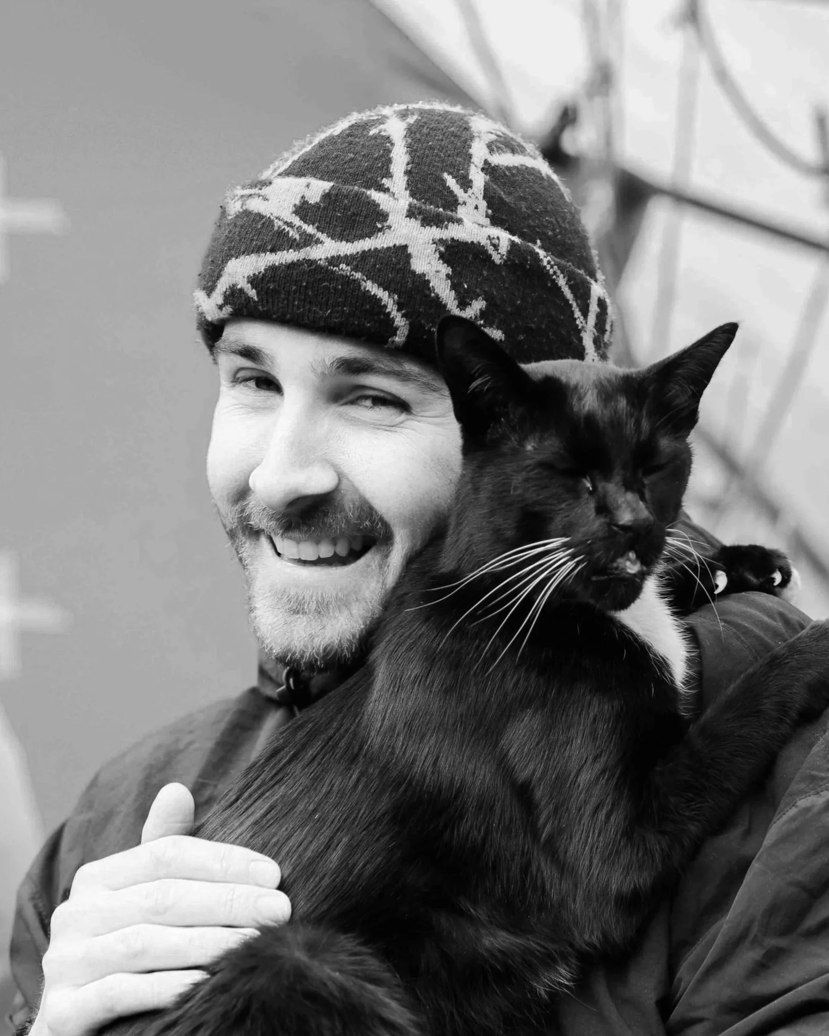 A smiling man wearing a patterned beanie, holding a black cat with white markings close to his face.