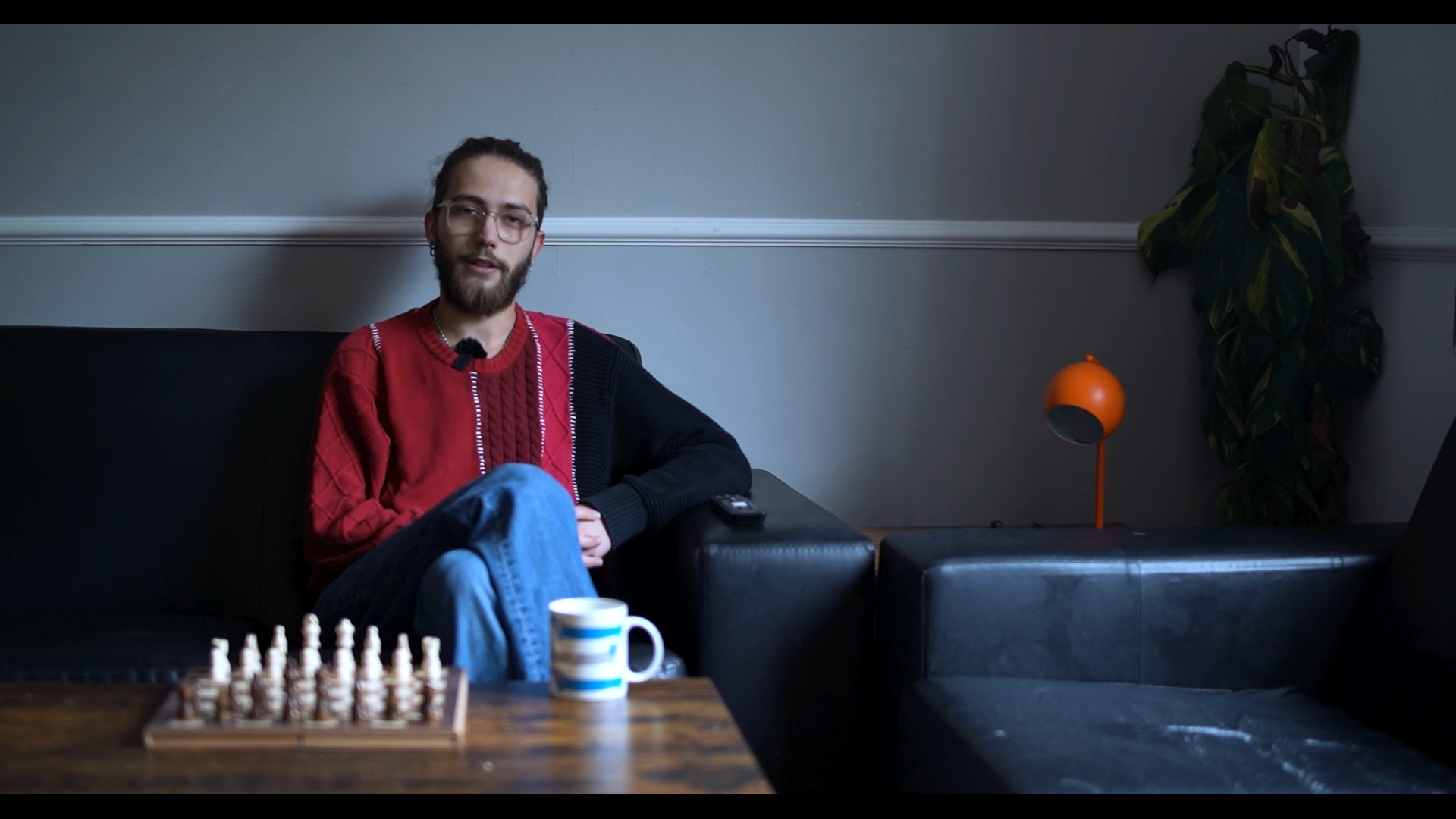 A man with glasses, a beard, and long hair tied back, sitting on a black leather couch in a dimly lit room, with a chessboard and a striped mug on a wooden table in front of him, and a tall green plant and an orange lamp in the background.