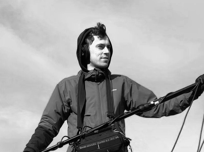 A young man with curly hair wearing a jacket and headphones, standing outdoors with a bicycle, looking to the side.