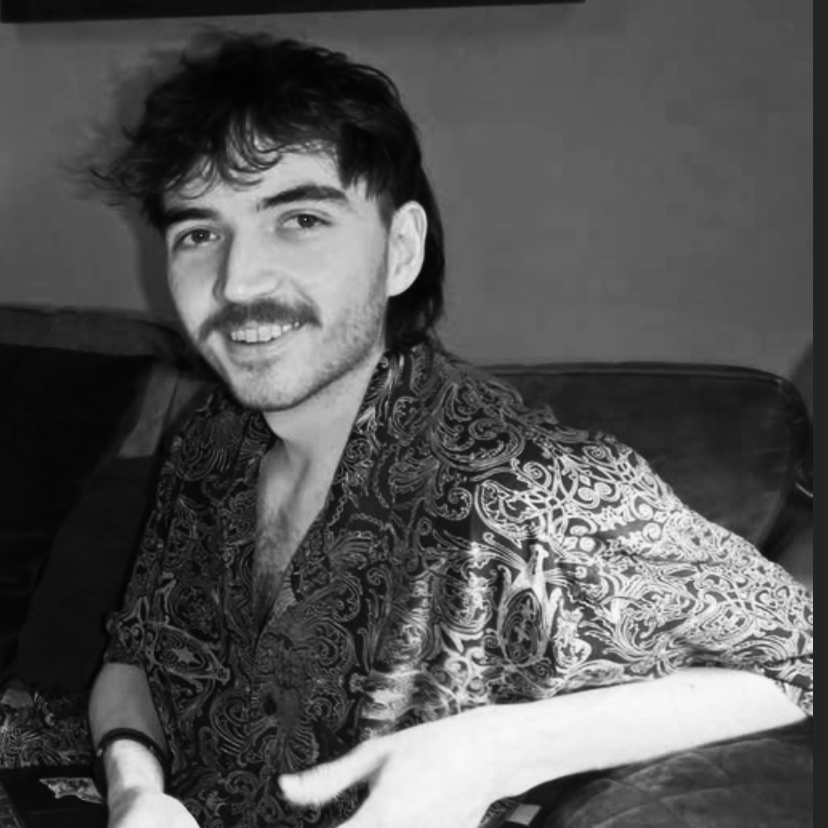 A young man with dark hair and a mustache smiling, sitting on a sofa, wearing a patterned shirt, in a black-and-white photo.
