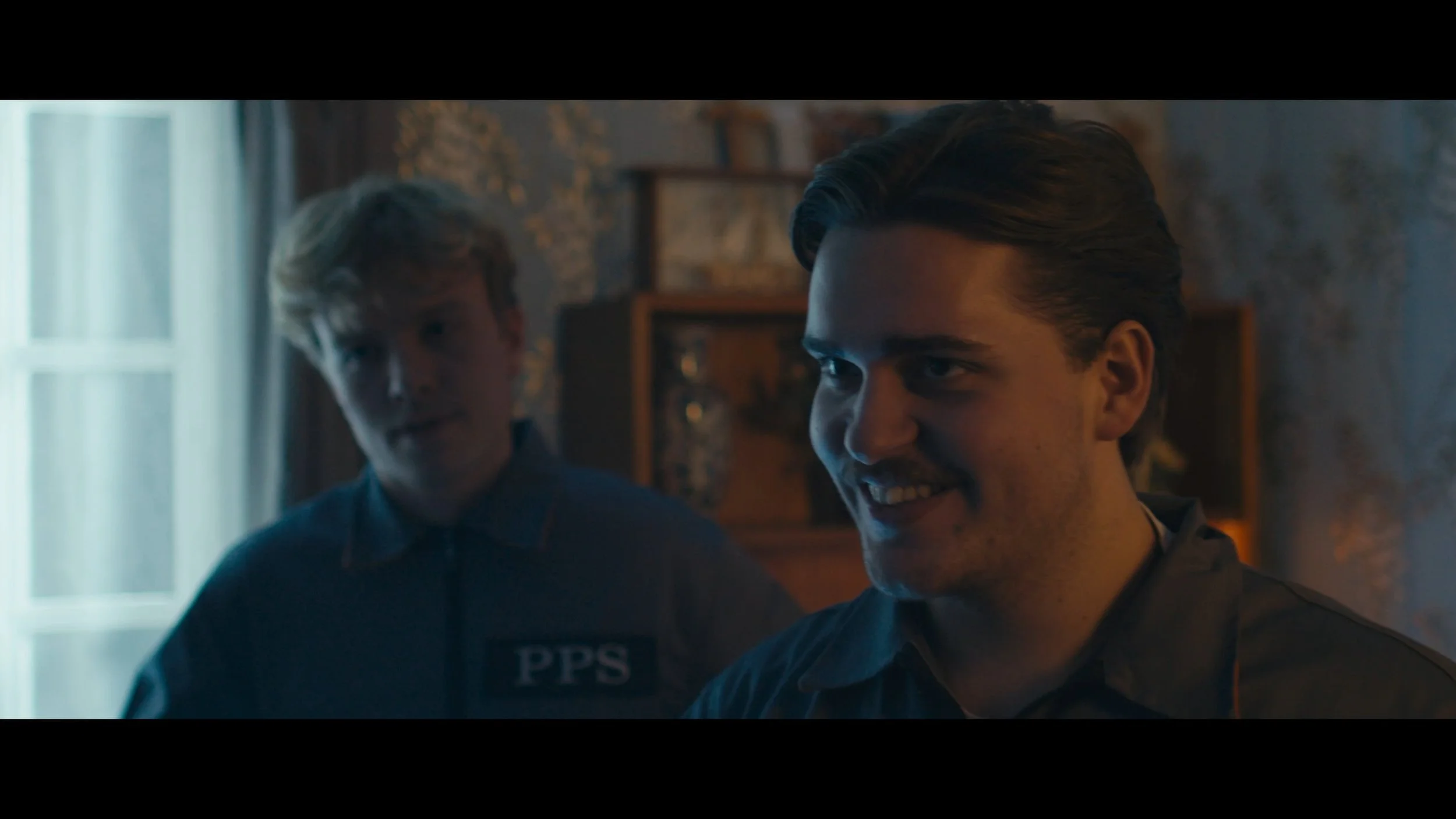 Two young men smiling indoors, one with blonde hair wearing a dark shirt with a patch saying 'PPS', the other with dark hair slicked back, in a room with a window and patterned wallpaper.