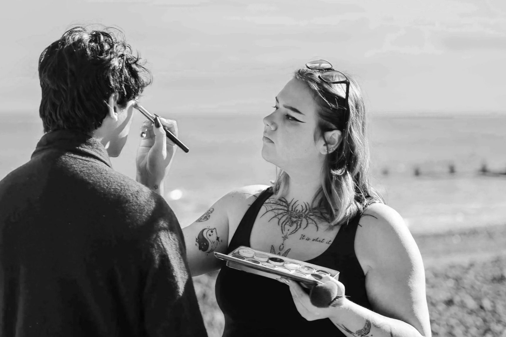 A woman with tattoos on her arm and chest getting her face painted by an artist on a beach with the ocean and sky in the background.