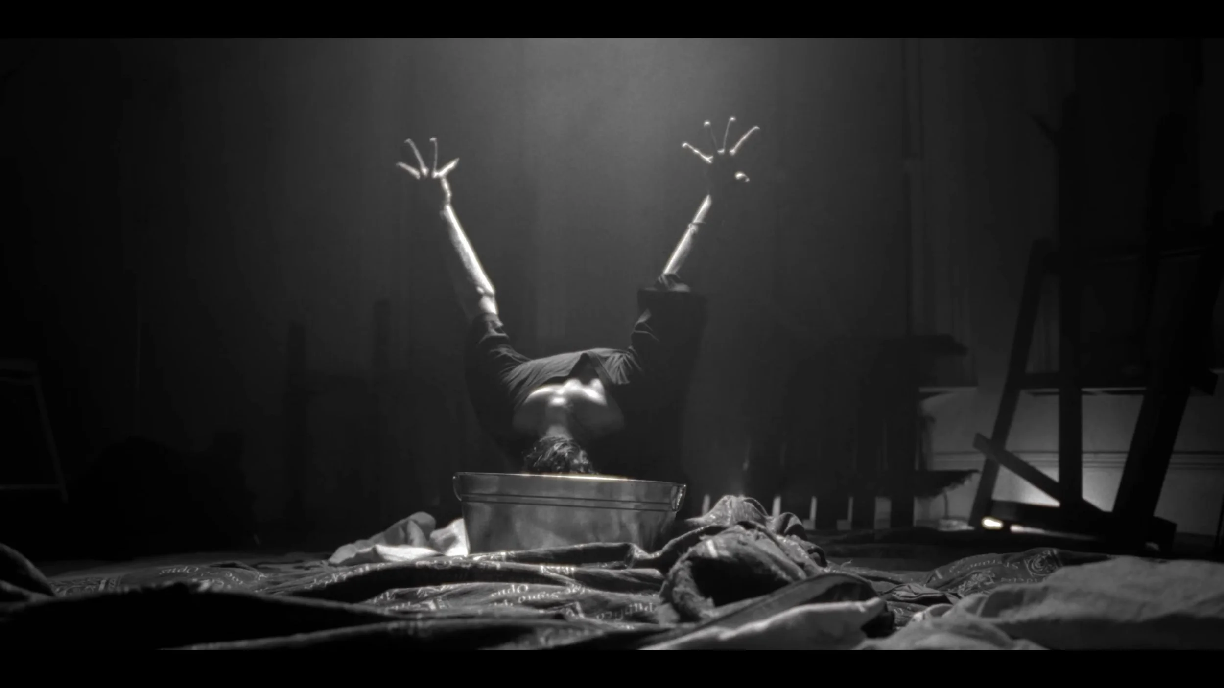A person with a head inside a metal basin, arms raised with fingers spread, in a dimly lit room with scattered objects and overturned furniture.