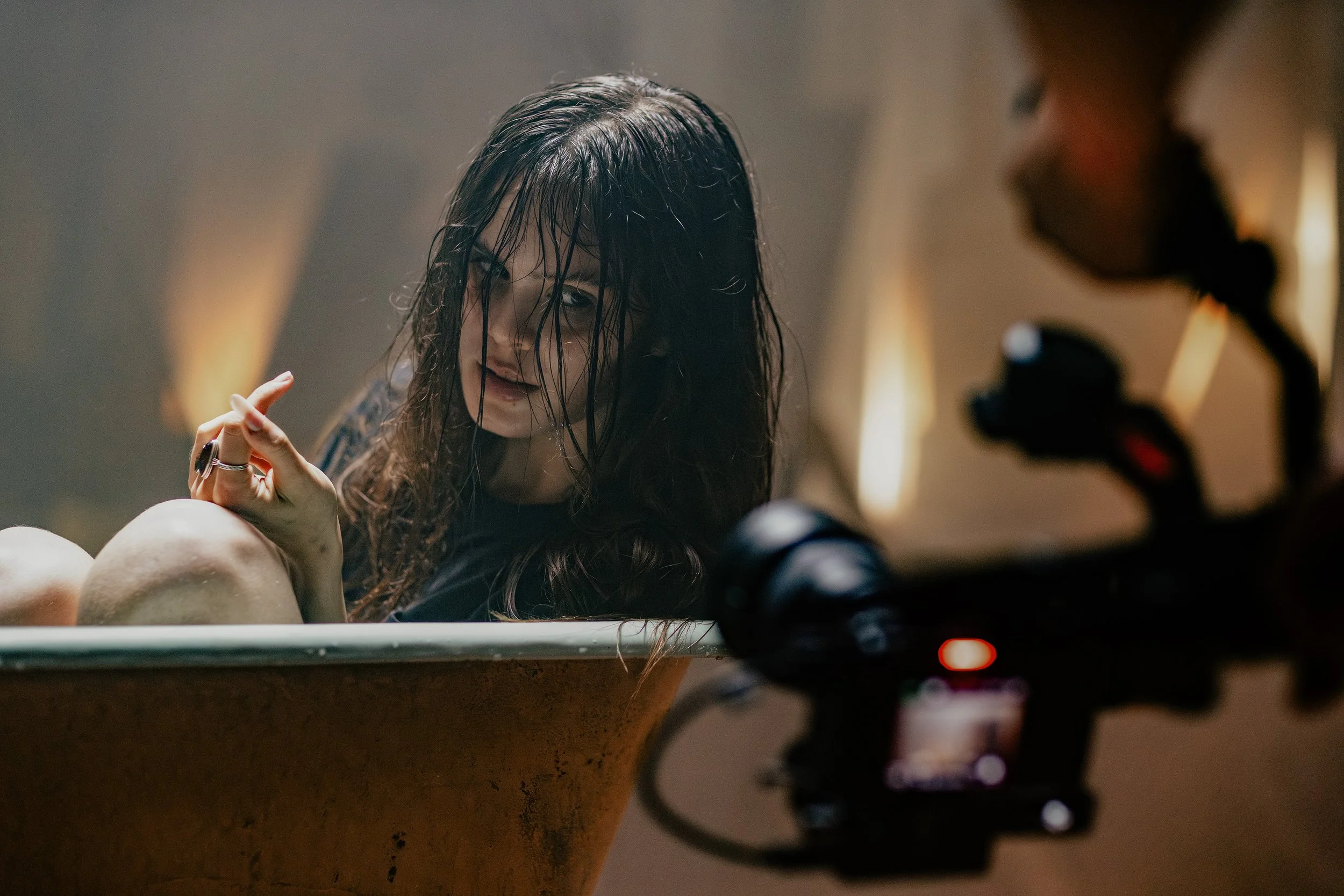 A woman with wet hair in a bathtub, looking at a camera, with a dark background and warm lighting. A camera on a gimbal is in front of her. Sofia Isella music video, "MUSE".
