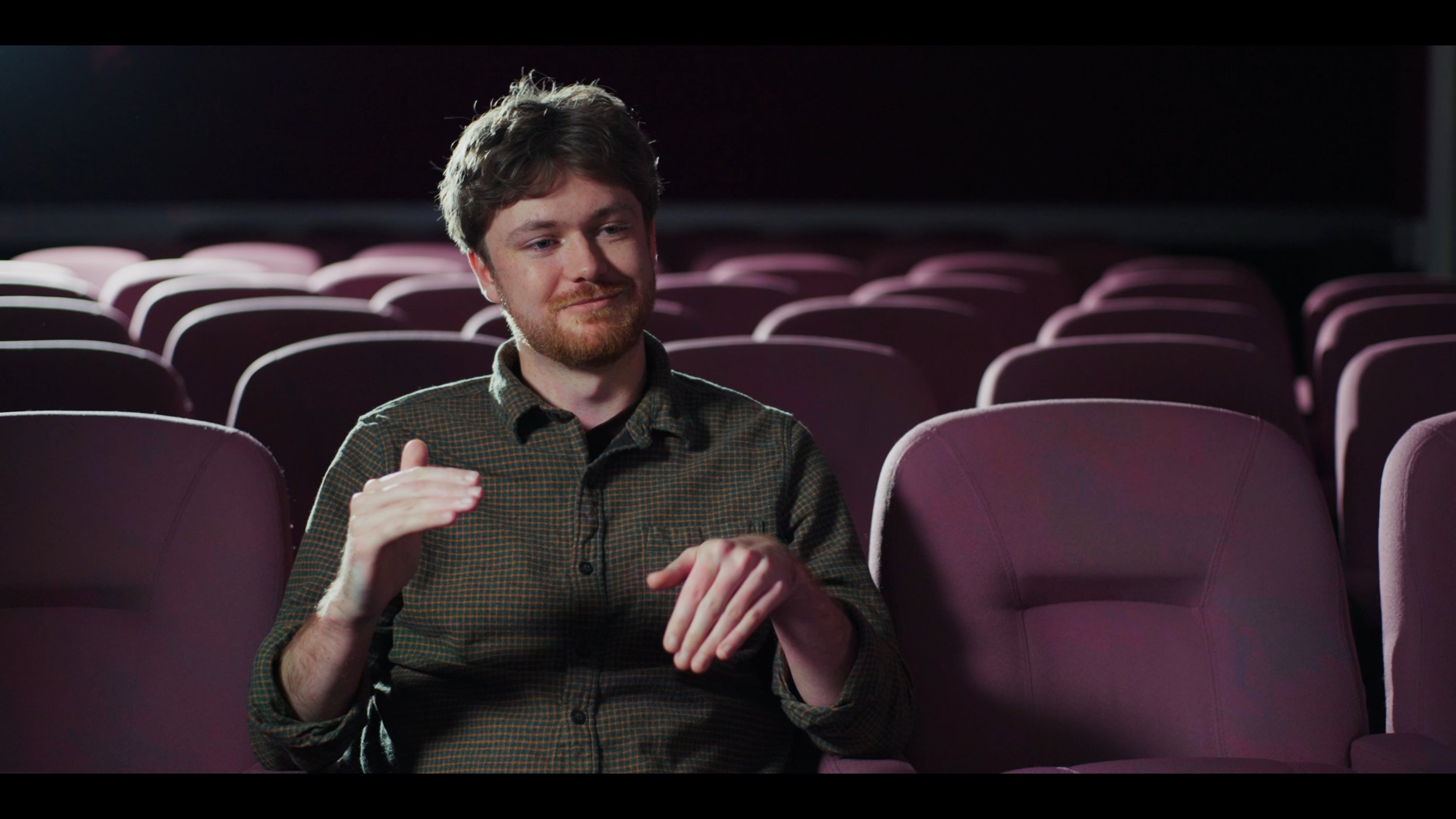 A man with brown hair and a beard sitting alone in an empty movie theater with maroon seats, gesturing with his hands.
