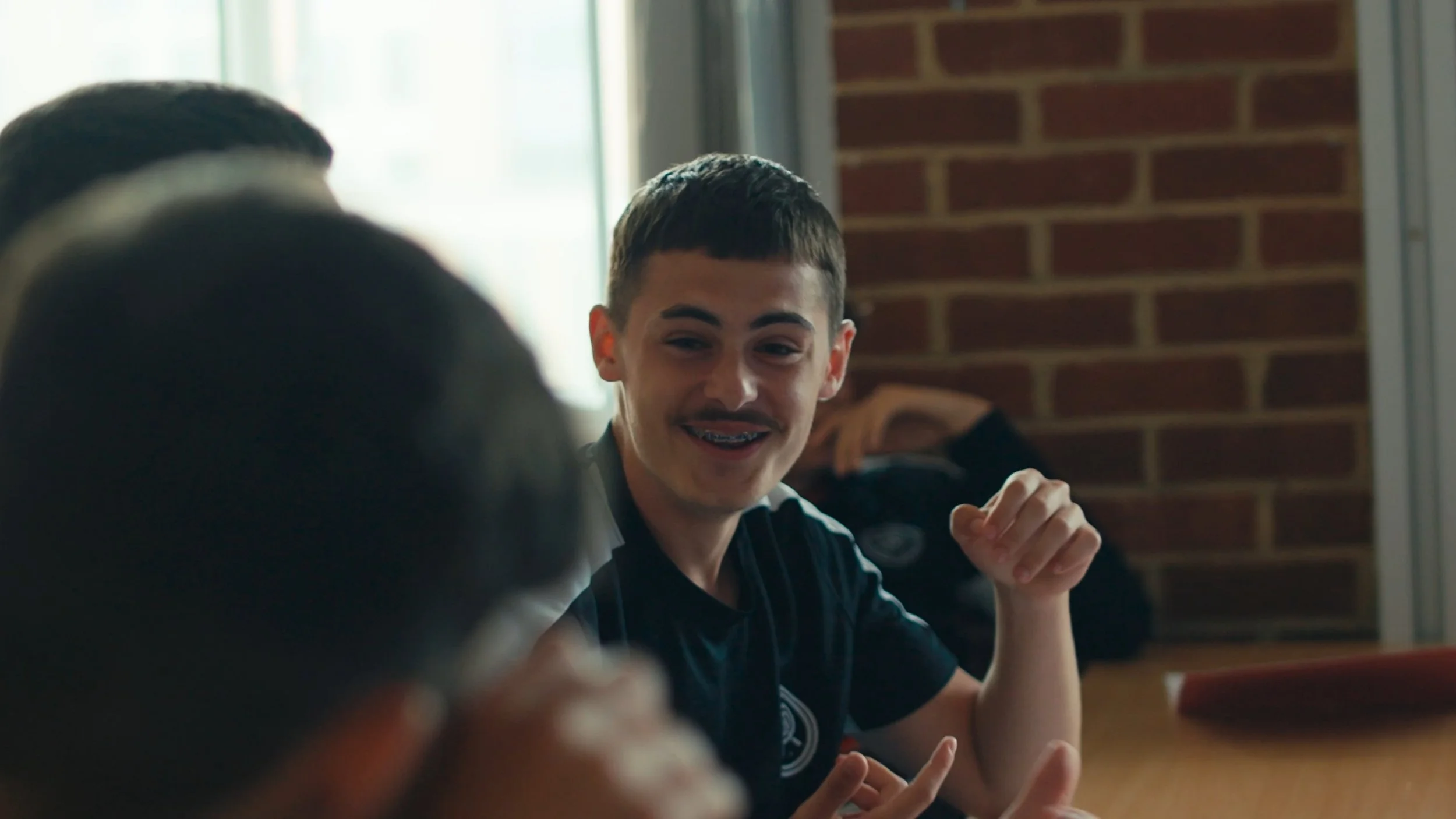 Young man with braces smiling and talking at a table indoors with other people.
