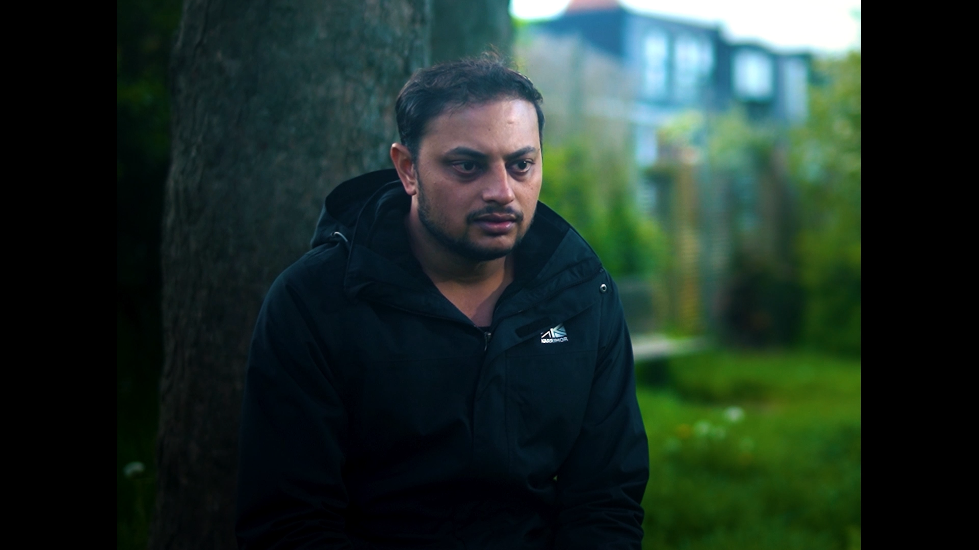 A man wearing a black jacket sitting outdoors near a tree with a contemplative expression, with a blurry background of greenery and buildings.