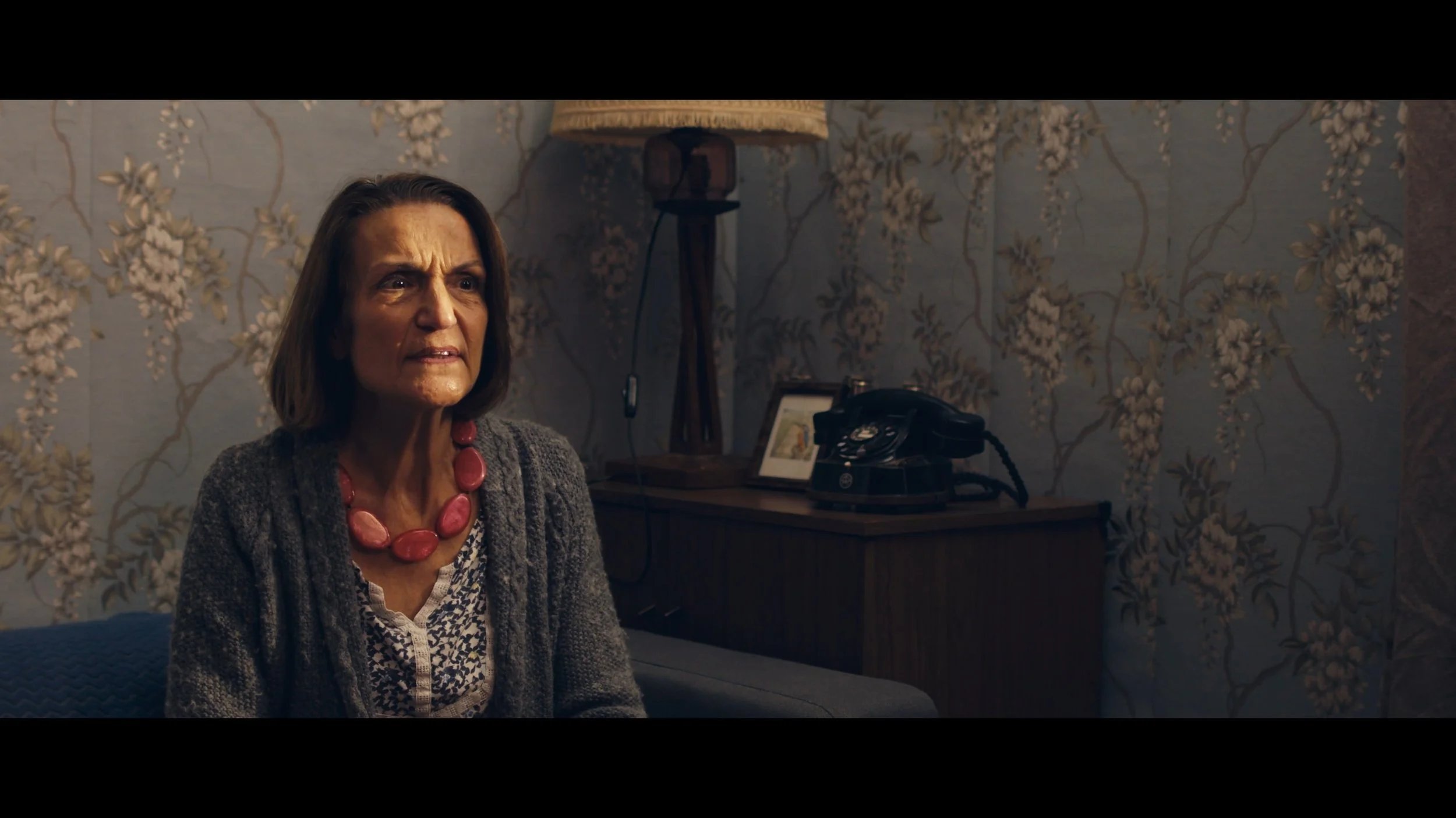 A woman with shoulder-length brown hair, wearing a gray cardigan and a pink beaded necklace, sits on a sofa in a room with floral wallpaper. She appears concerned or upset. There is a wooden side table behind her with a black rotary phone, a small framed picture, and a wooden lamp with a beige shade.