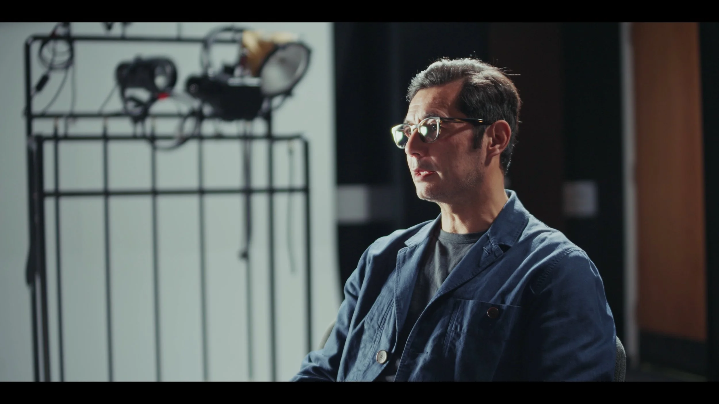 A middle-aged man with glasses and dark hair, wearing a denim jacket, sitting in a room with a black metal wire rack and headphones in the background.