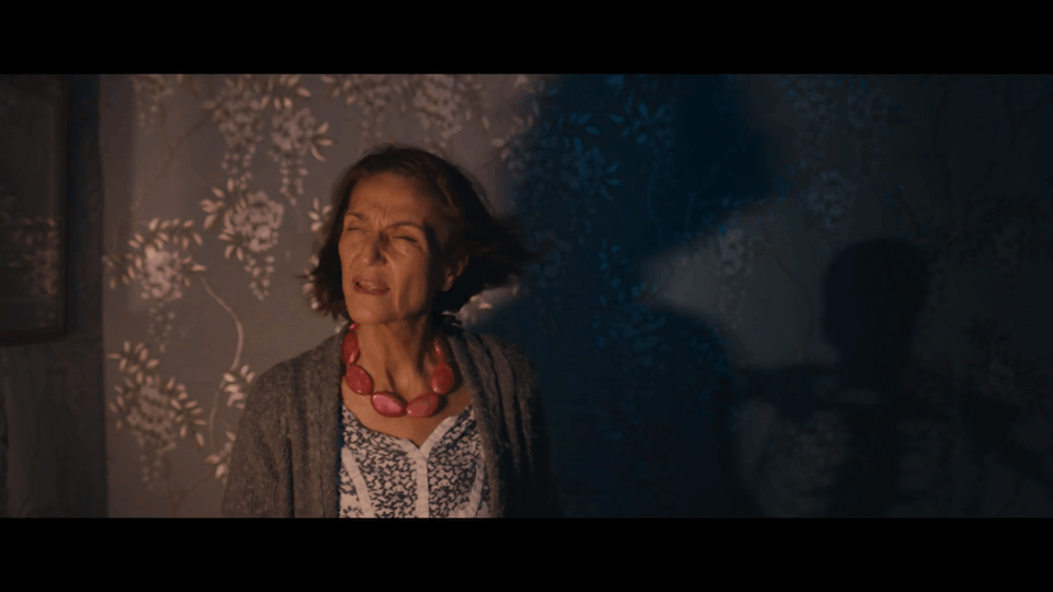 An elderly woman with short brown hair is standing in a dimly lit room with floral wallpaper, appearing distressed or alarmed.