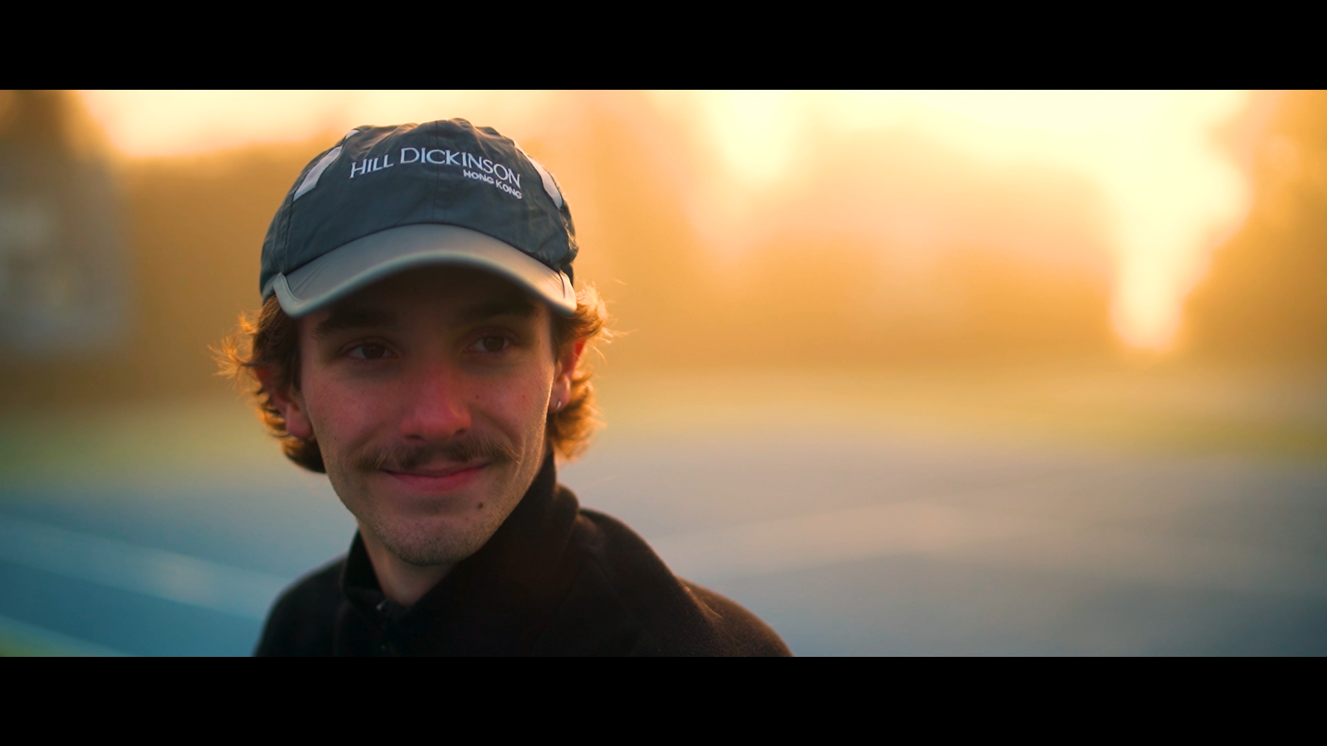 Close-up of a young man with light brown hair, mustache, wearing a black jacket and a dark blue baseball cap with grey brim, standing outdoors during sunset with blurred background of trees and sky.