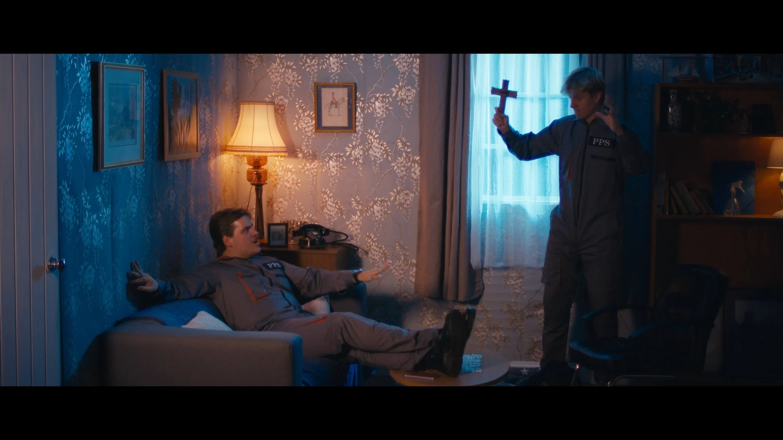 Two young men in security uniform in a room with floral wallpaper; one is lying on a couch with arms outstretched, the other is standing with a red cross, all creating a tense scene.