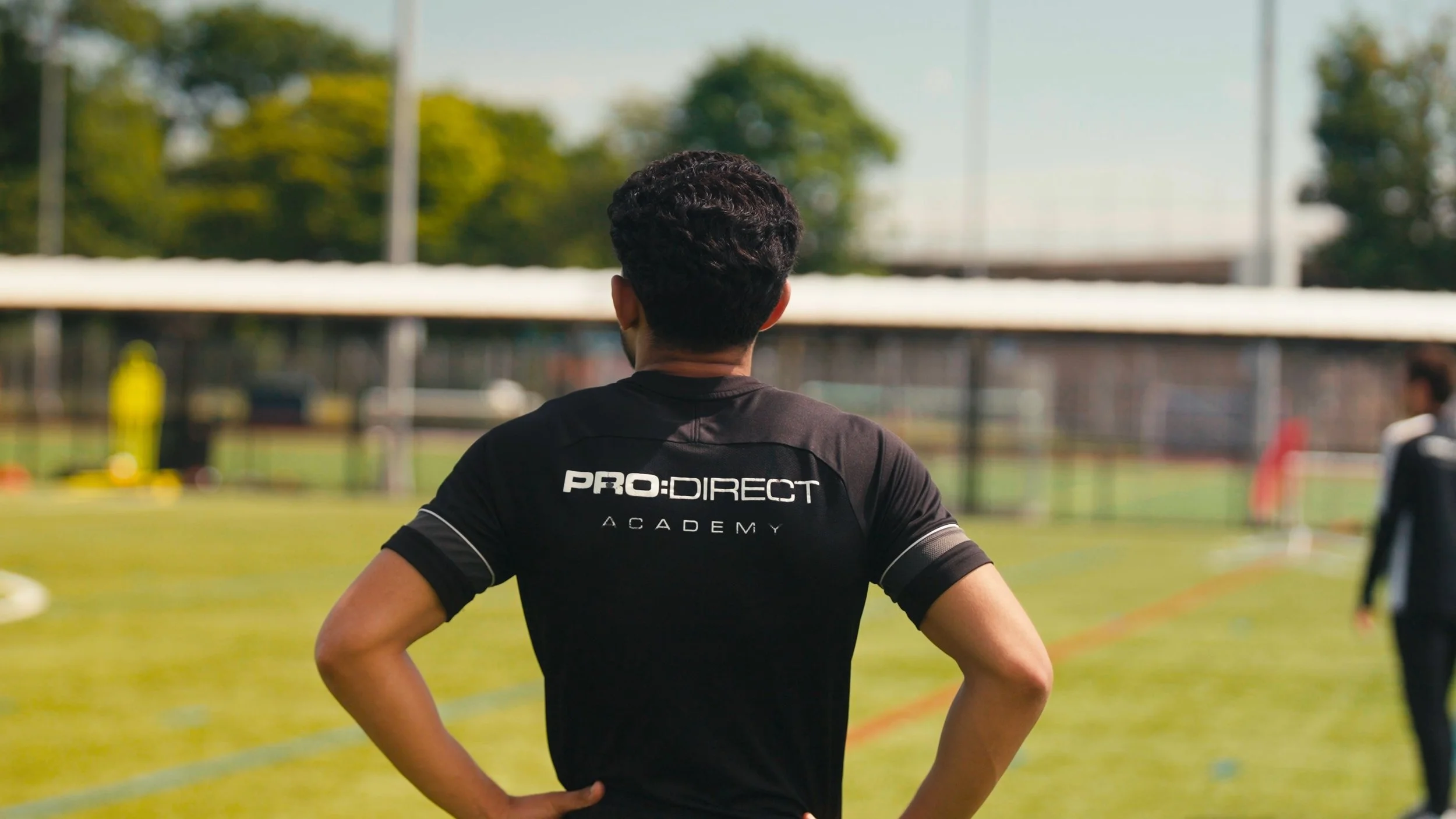A person with dark curly hair wearing a black T-shirt with 'PRO: DIRECT ACADEMY' written on the back, standing on a sports field with their hands on their hips, facing away from the camera. The background shows trees and a goalpost on the field.