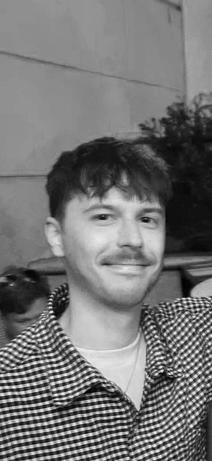 A young man with messy dark hair, wearing a hoodie with a logo on the front, standing indoors against a plain wall.