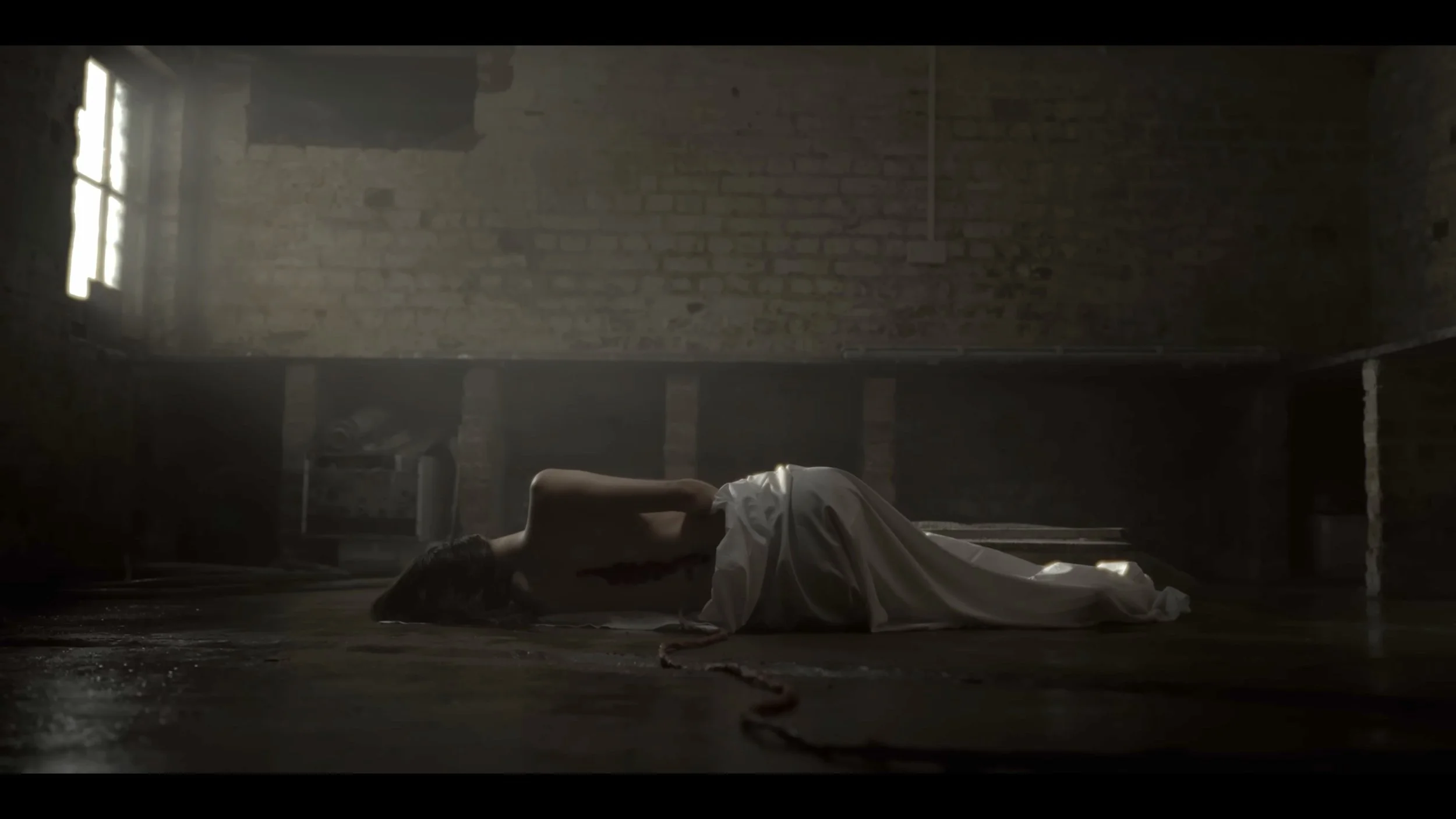 A woman lies on the floor in an unfinished brick building, partially covered with a white sheet, with a rope beside her and soft sunlight coming through a small window.
