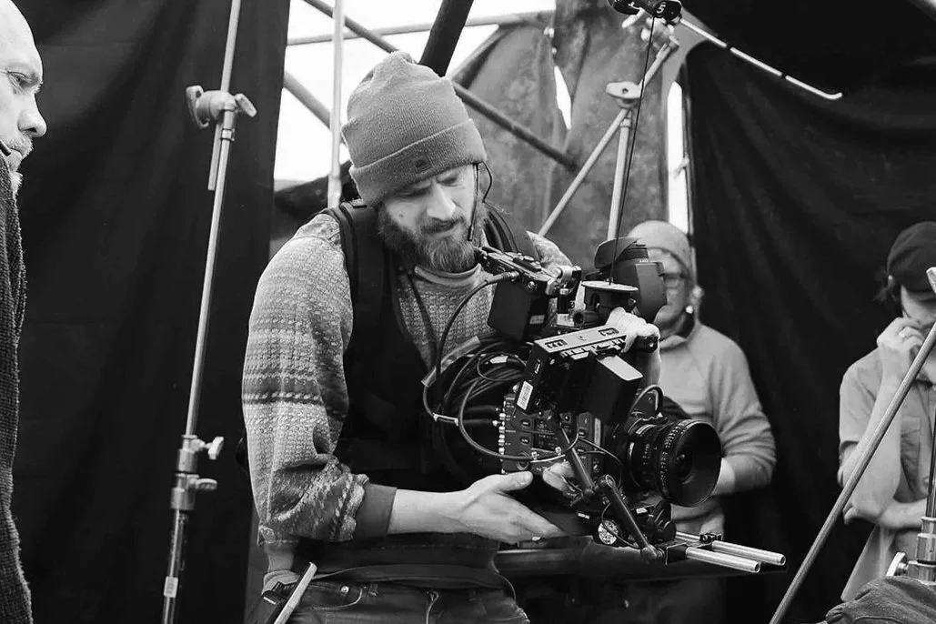A man with a beanie and beard operating a professional film camera on a set with crew members in the background.