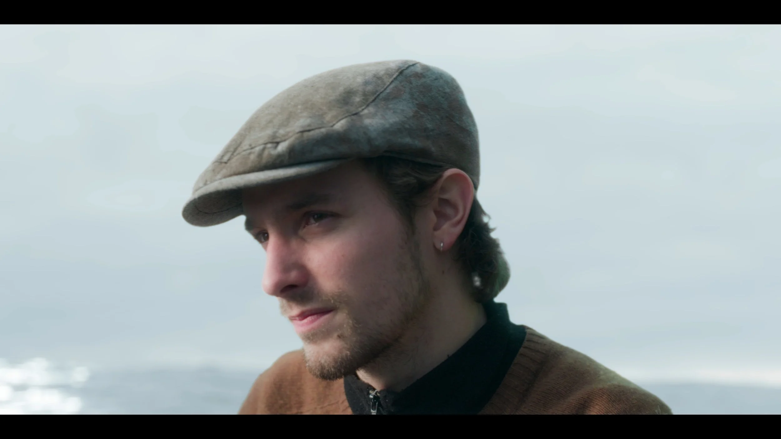Close-up of a young man with a brown jacket, a flat cap, a beard, and a small earring, outdoors with a blurred ocean background.
