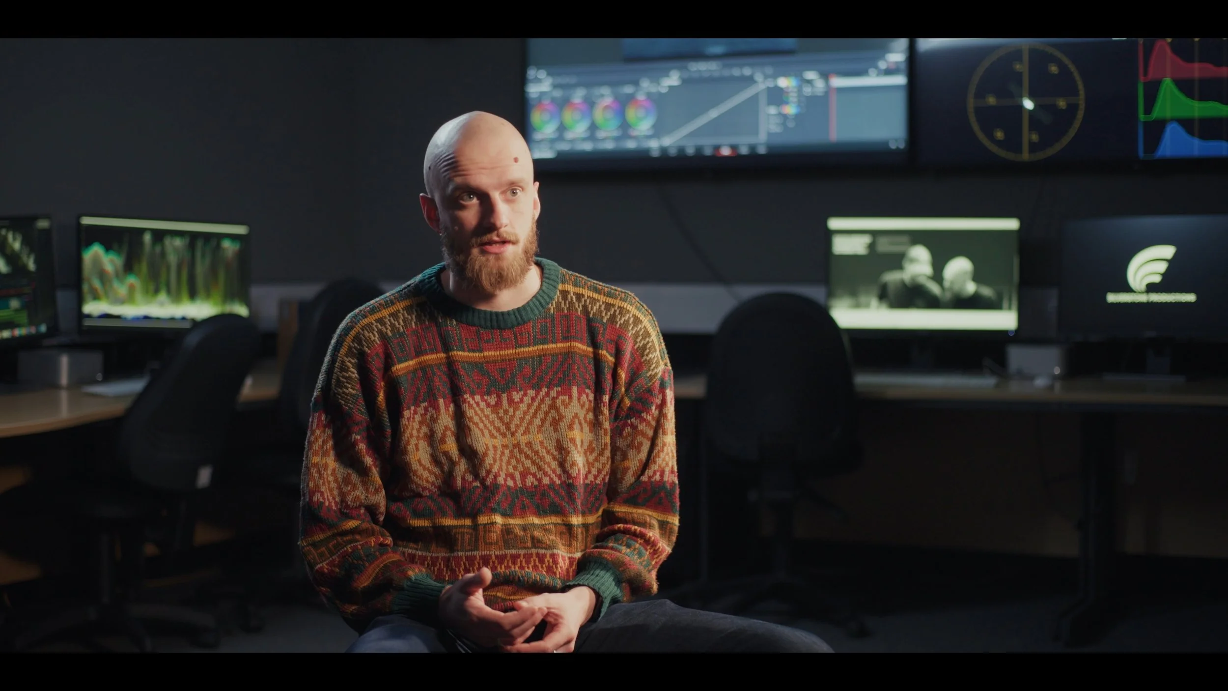 Man with a beard wearing a multicolored patterned sweater seated in a dark room with multiple computer monitors displaying graphs and images in the background.