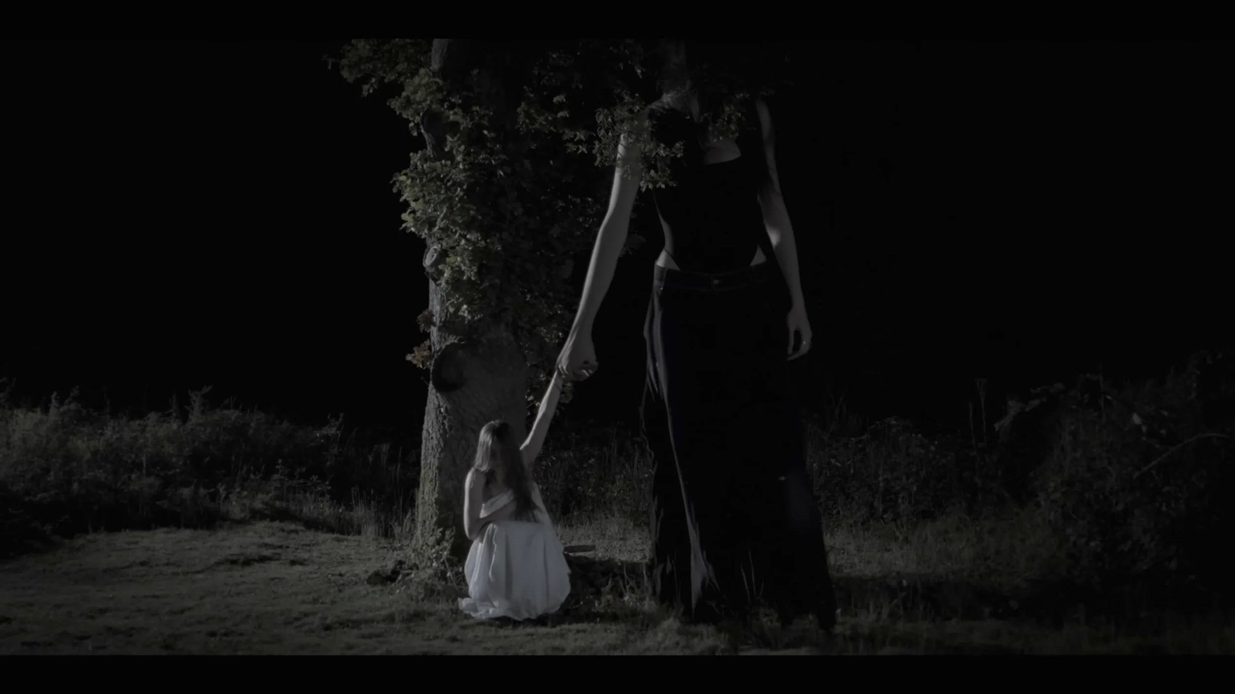 A woman in a long black dress is holding the hand of a young girl in a white dress under a tree at night in a dark outdoor setting.