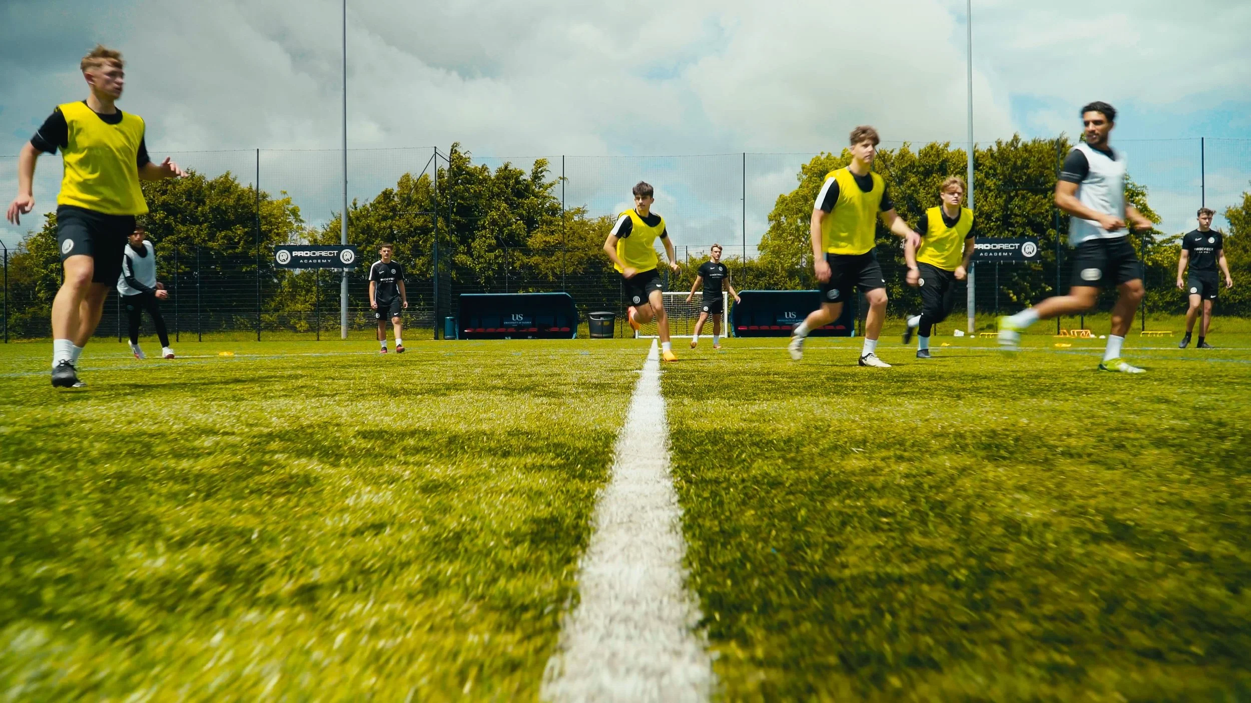 Young men playing soccer on a grass field during practice, with some wearing yellow training vests.