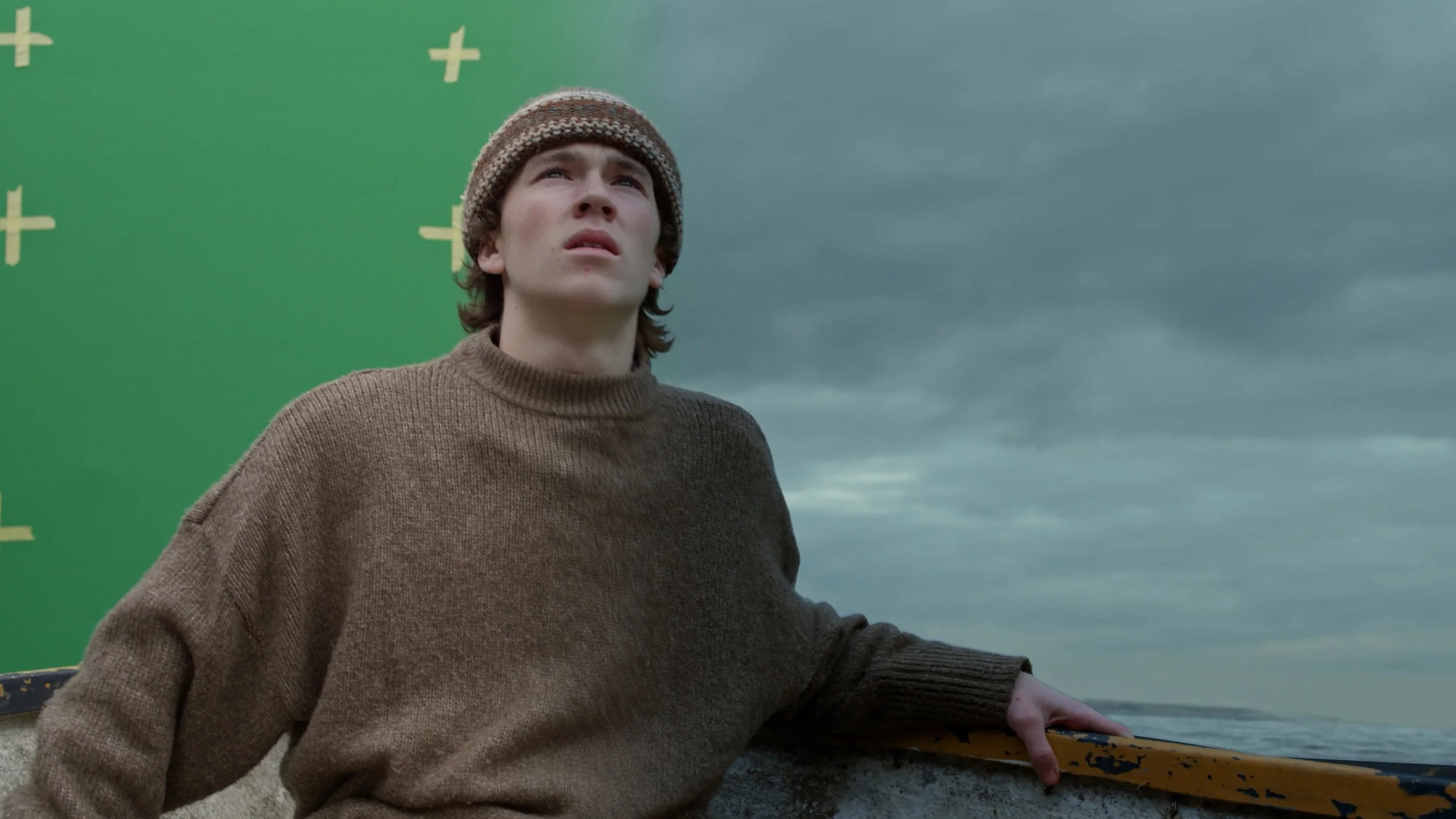 A young man in a brown sweater and knit cap looks up with a concerned expression, standing outdoors beside an old yellow boat, with a green screen on one side and a cloudy sky in the background. Visual Effects, VFX. Blender. Nuke. Compositing.