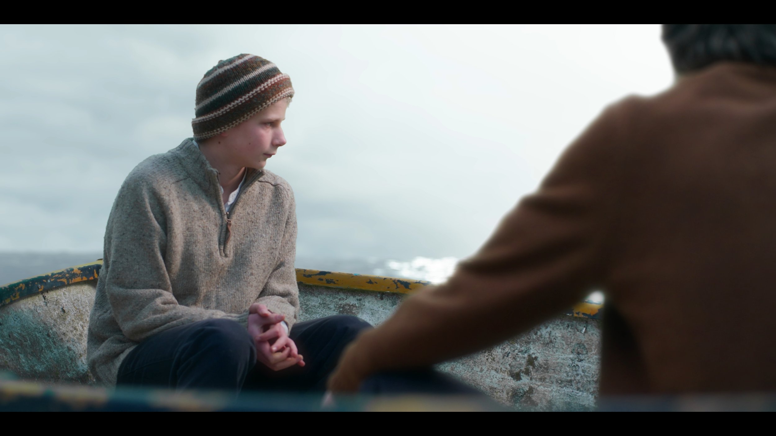 A young boy in a beige sweater and striped beanie sitting in a small boat, looking down with a serious expression, while an adult's arm reaches toward him over the side of the boat, with a cloudy sky and water in the background.